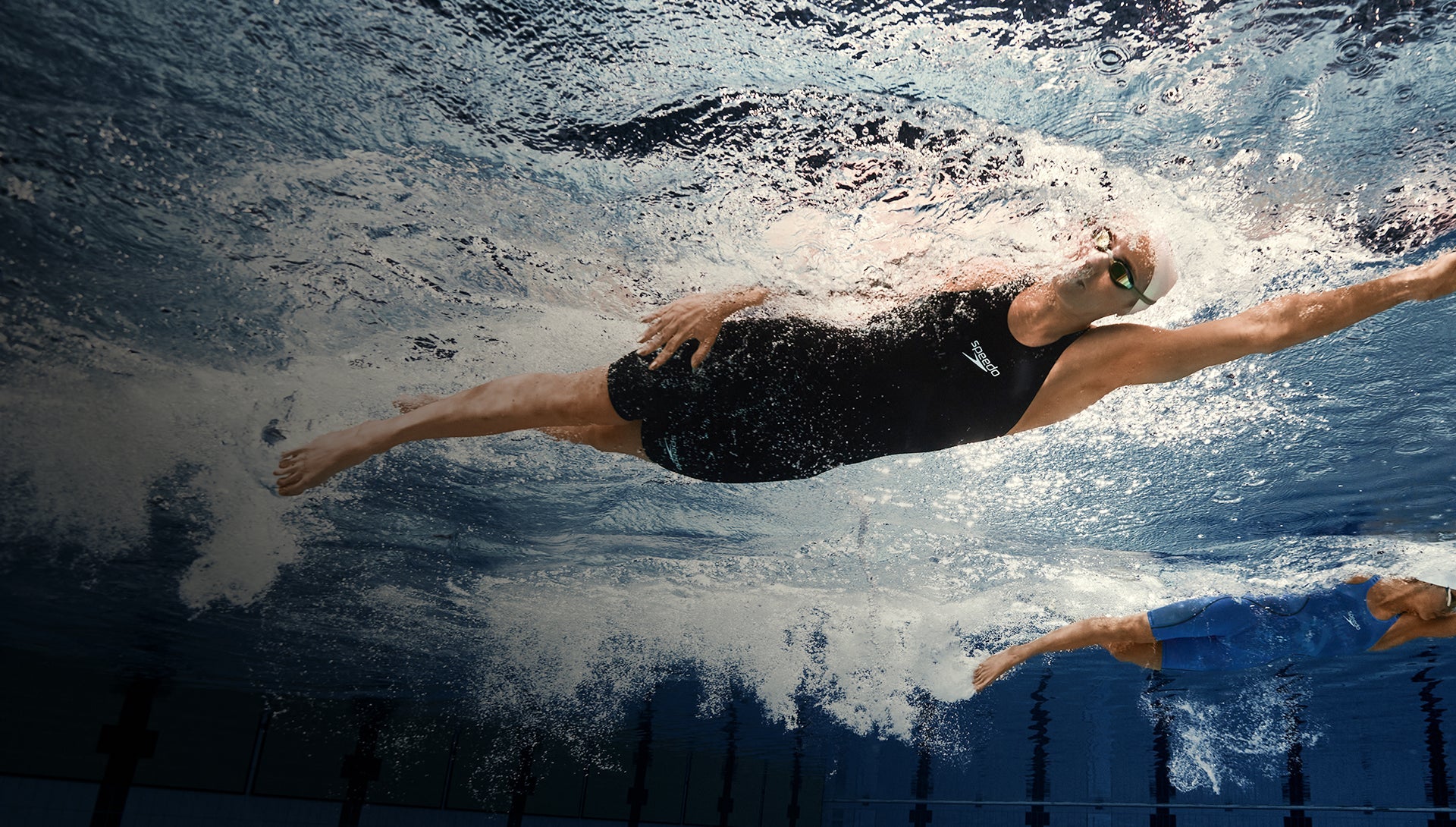Swimmer gliding underwater in a pool with another swimmer behind.