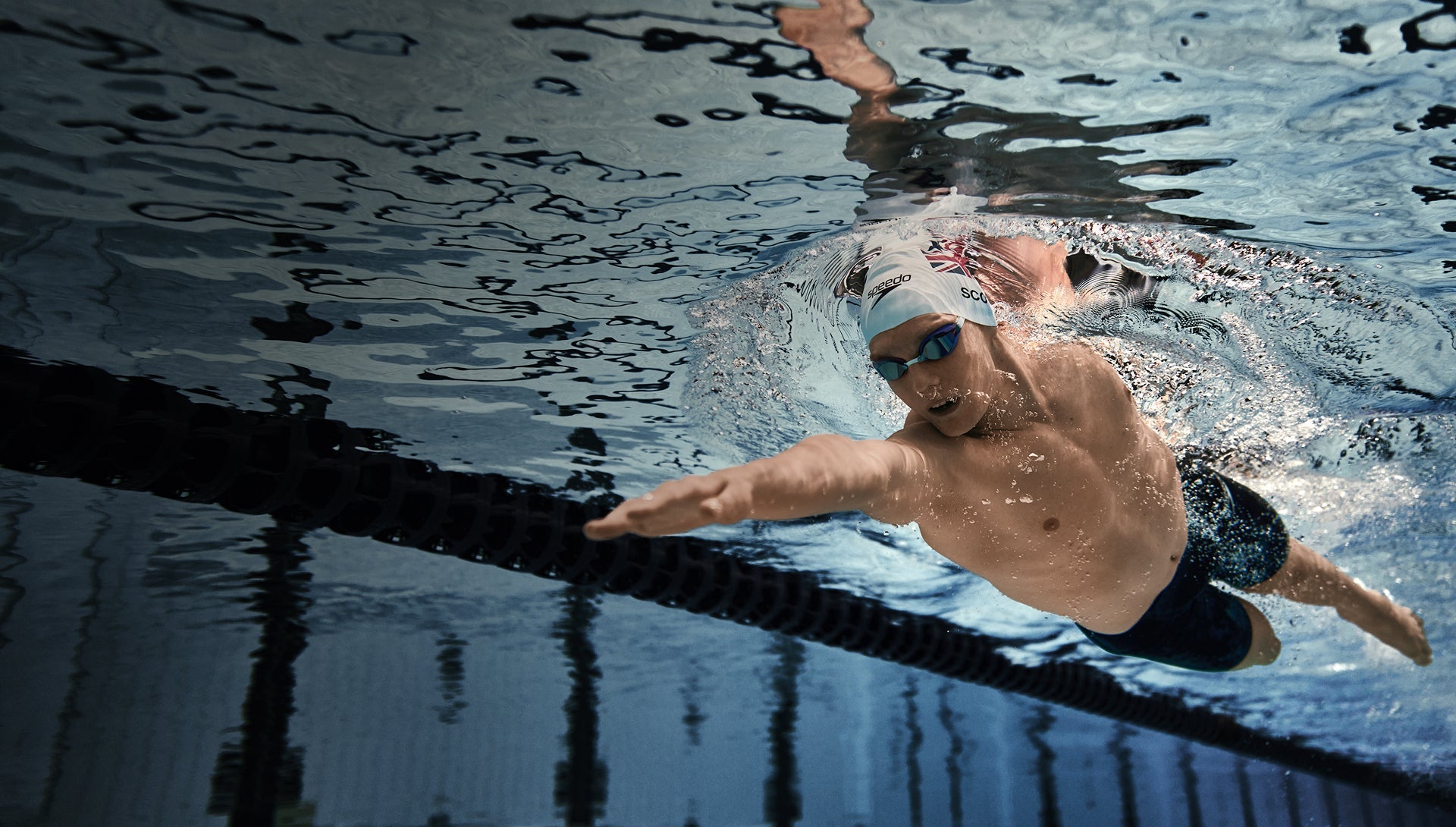 Swimmer reaching forward underwater in a pool, wearing a cap and goggles.