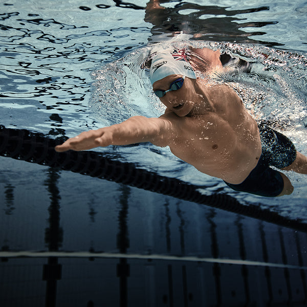 Swimmer reaching forward underwater in a pool, wearing a cap and goggles.