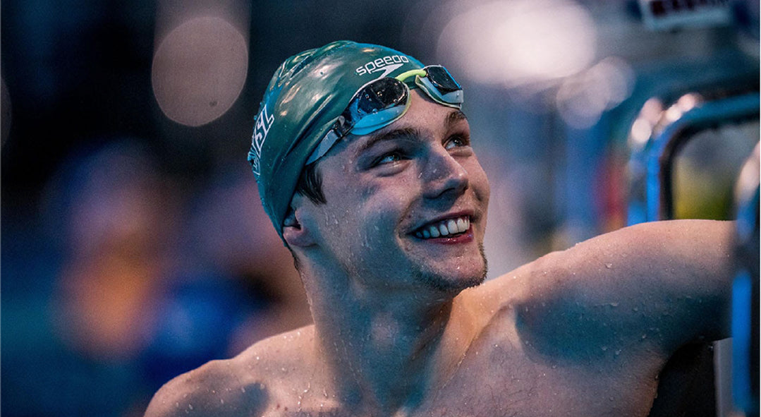 Smiling swimmer in a green cap and goggles by the pool.