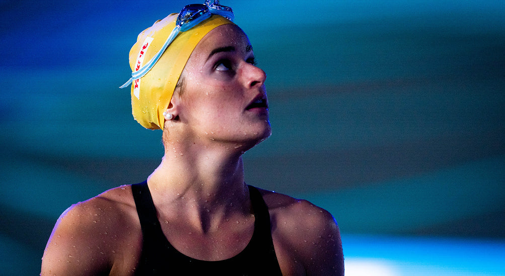 Close-up of a swimmer in a yellow cap and goggles looking upward, water droplets on her face.