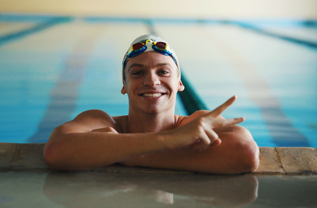Smiling swimmer resting at the pool edge, wearing goggles and making a peace sign.