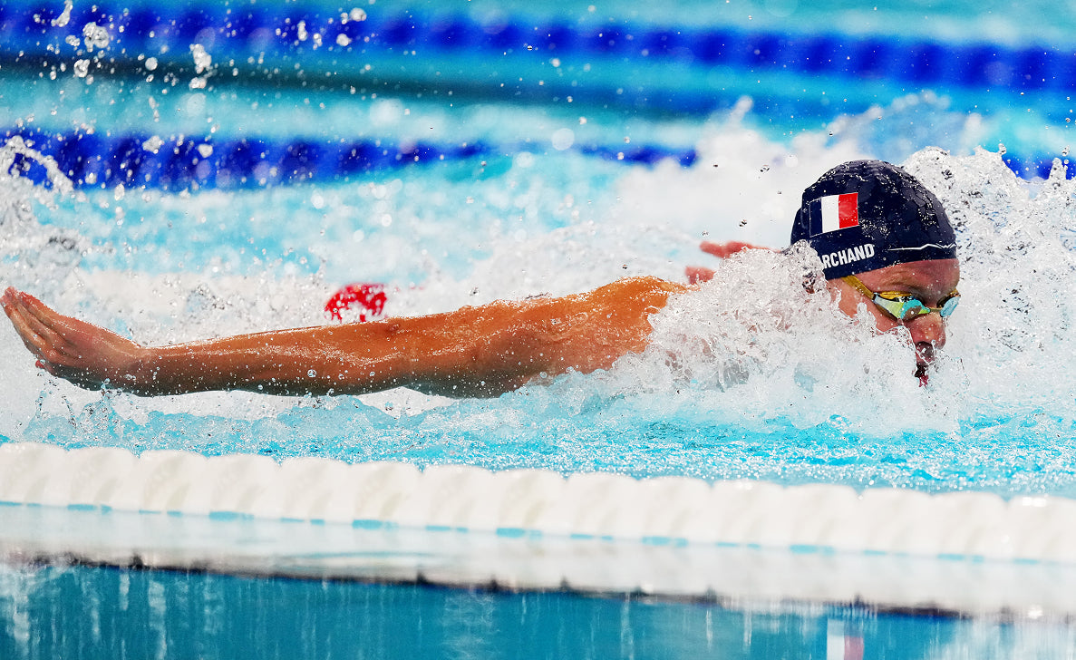Swimmer doing butterfly stroke in a pool, splashing water, wearing a cap and goggles.